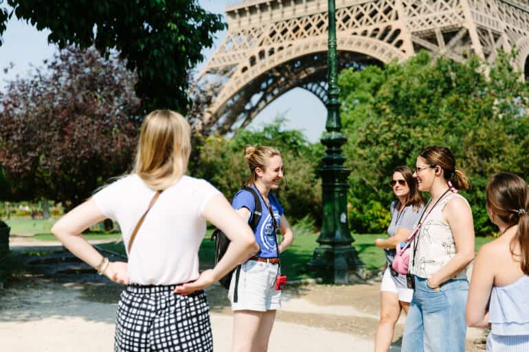 Tour guide smiles and shares a laugh with a group of people with the base of the Eiffel Tower in the background.