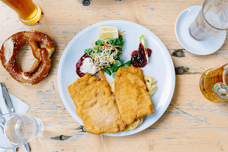 Flatlay of a traditional German spread at a restaurant including a plate of schnitzel, a pretzel, and beer.