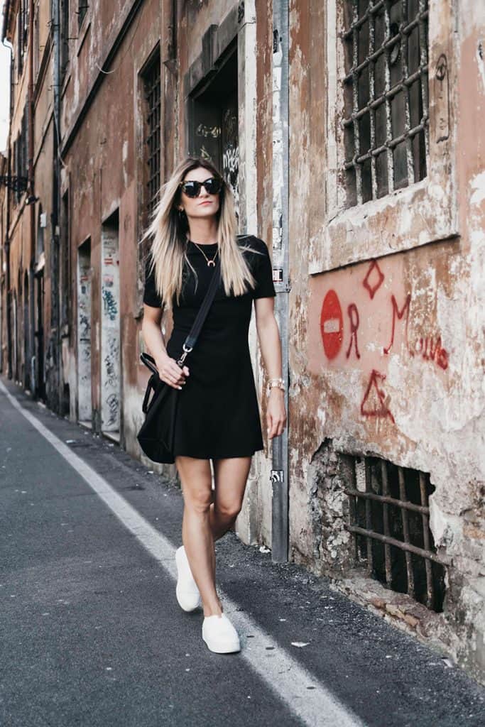 A woman wearing a black dress, sunglasses, and white trainer walks along the street in Rome, Italy.