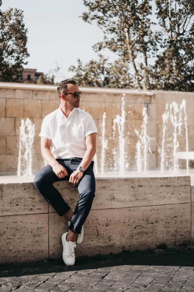 A stylish man wearing a crisp white button up and white sneakers pauses next to a fountain in Rome, Italy.