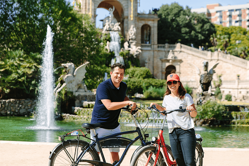 Two people smile next to bicycles in front of the fountain of The Parc de la Ciutadella in Barcelona, Spain