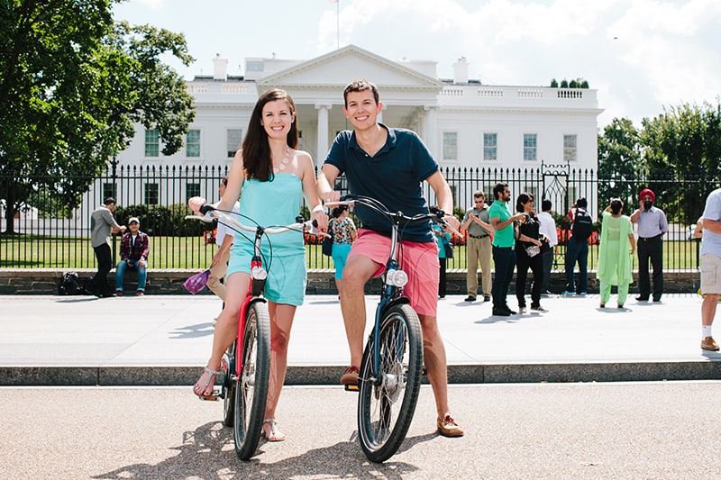 Two people pose on bikes in front of the White House in Washington, D.C.