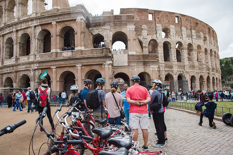 A group of people on a bicycle tour stand outside the Roman Colosseum and listen to their tour guide