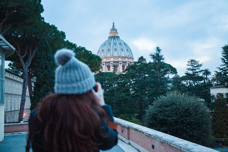 Woman wearing a beanie takes a photo of the Vatican Dome from a distance