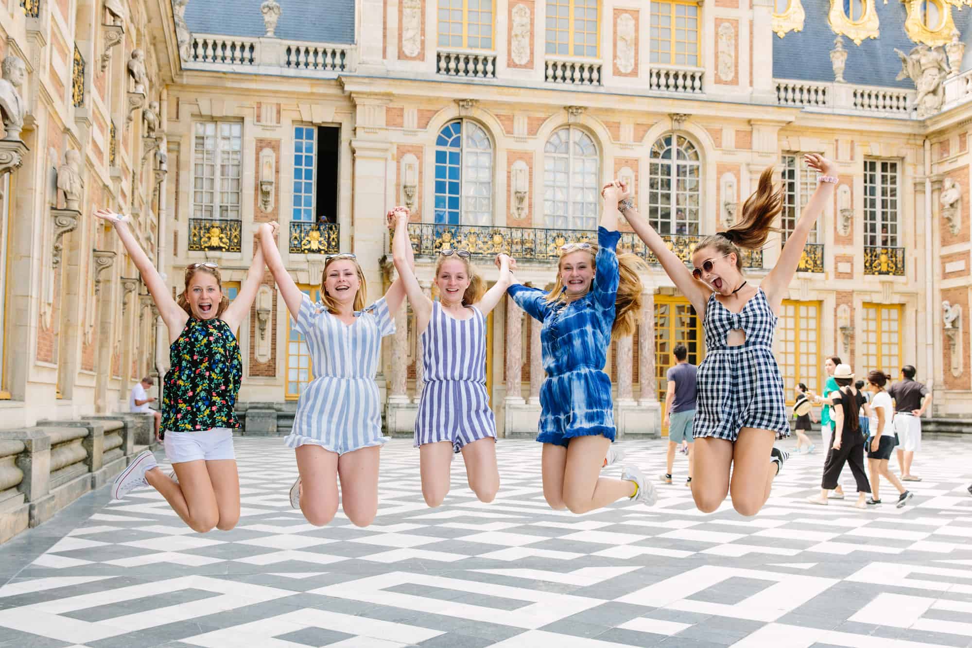 Five friends hold hands and jump in front of the Versailles Palace.