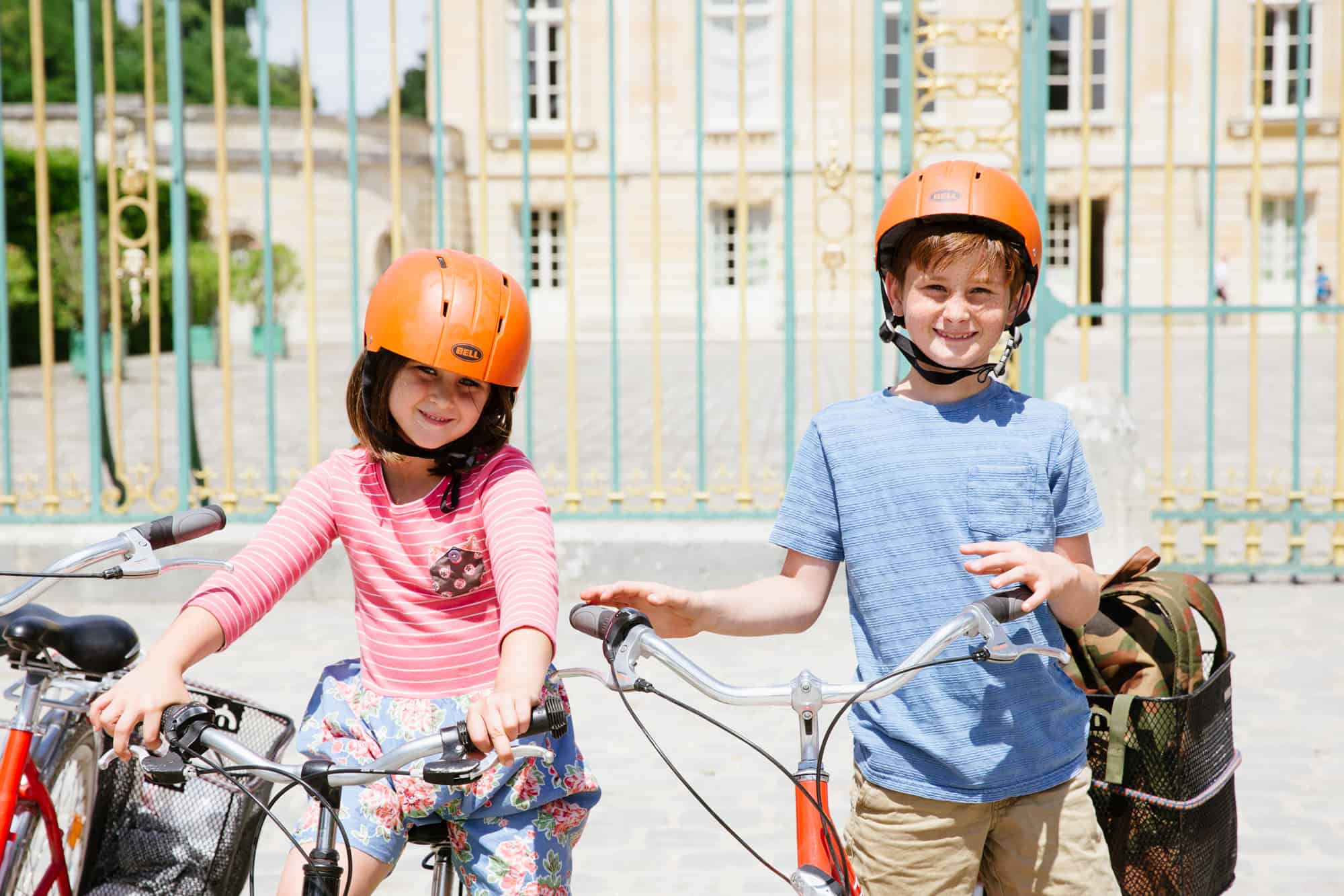 Two children wearing orange helmets pose for a photo with their bikes in front of the Petit Trianon in Versailles.