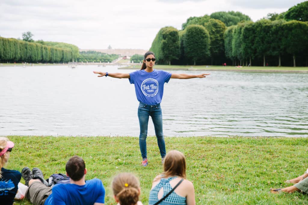 A guide in a blue t-shirt explaining the chateau of Versailles to a ground while they're seated in the grass along the Grand Canal