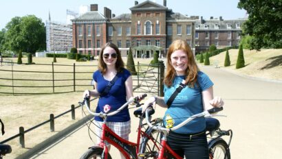 Two women pose with their bikes in front of Kensington Palace in London, England