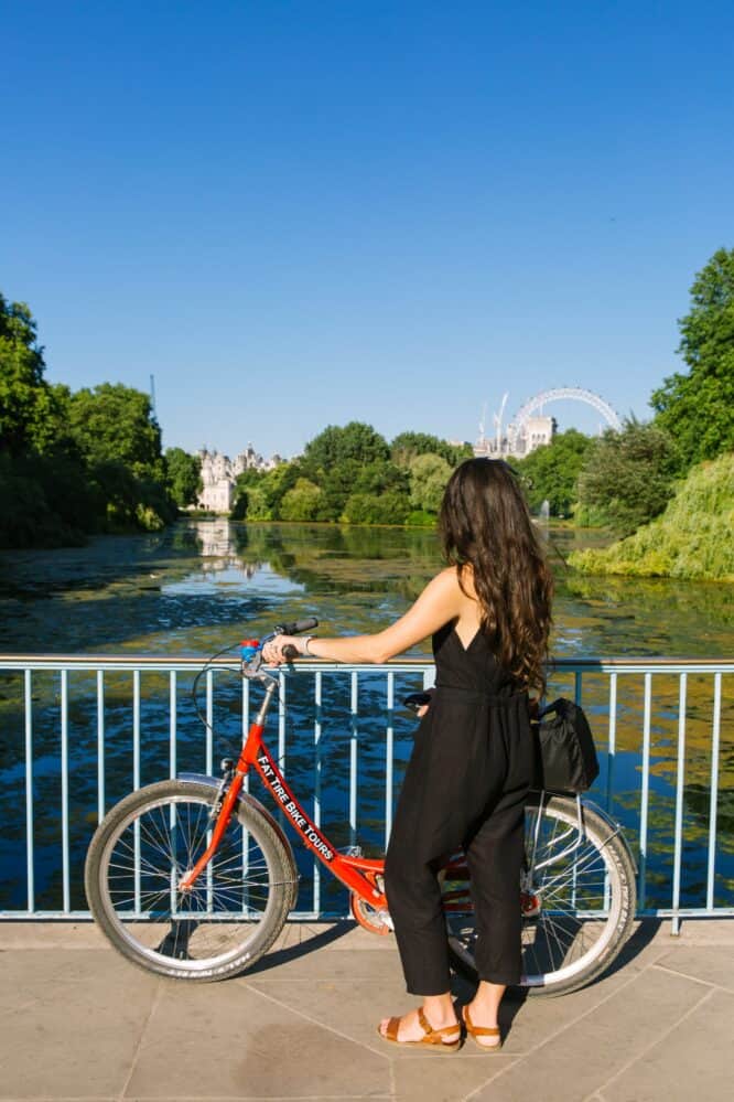 woman standing next to her red bicycle looking at London from a bridge over water