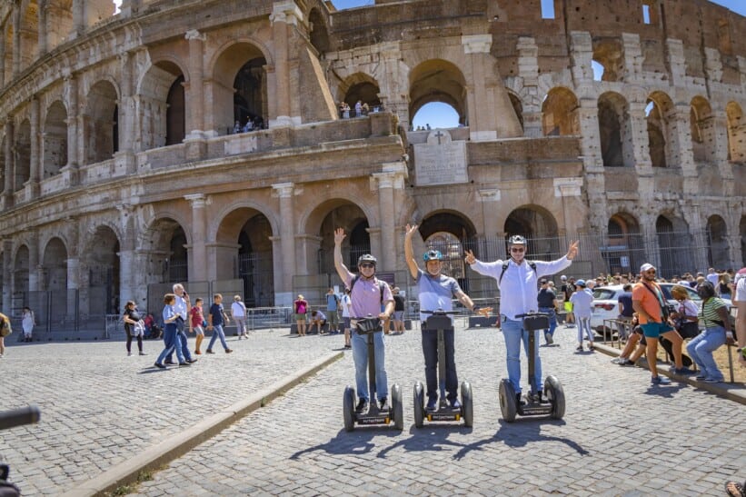 A group of Segways pose for a photo with their hands in the air in front of the Colosseum in Rome, Italy
