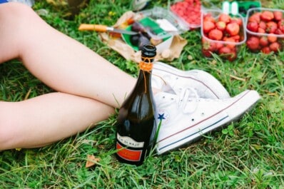 A bottle of bubbly leaning against a women's leg as she's sitting in the grass