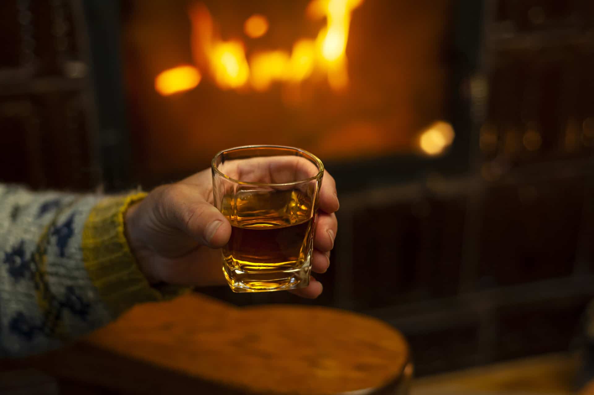 A man holds a glass of whiskey in front of a fireplace