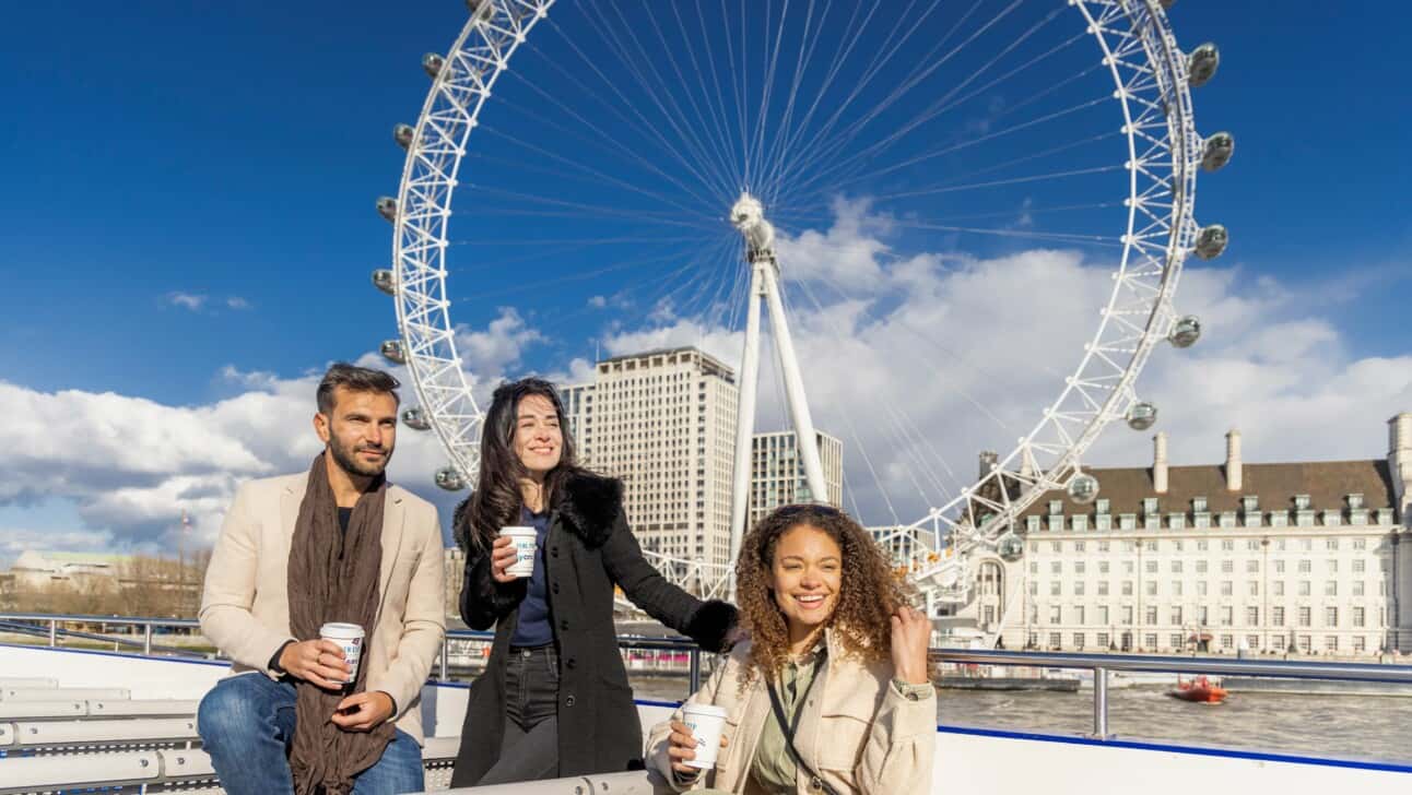 Friends pose for a photo with warm drinks in hand in front of the London Eye
