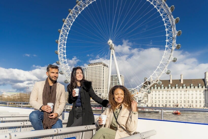 Friends pose for a photo with warm drinks in hand in front of the London Eye