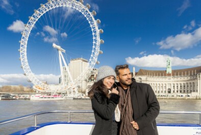 A couple poses for a photo in front of the London Eye while on a River Thames cruise
