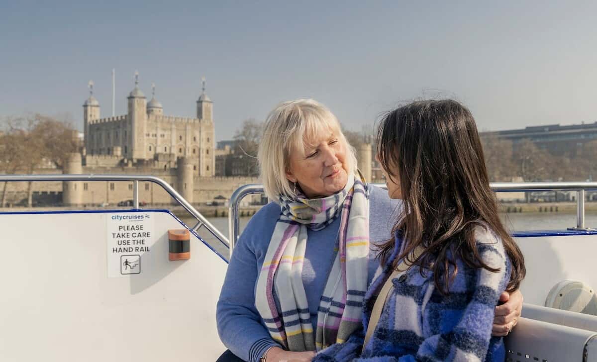 Two women have a conversation on a boat with the Tower of London in the background.