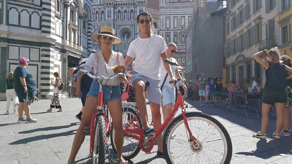 a couple poses with their bikes and a small dog in front of the Duomo in Florence, Italy