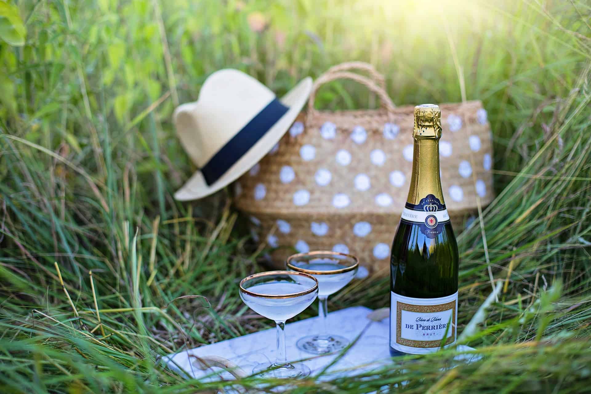 A bottle of champagne, two glasses, and a picnic basket with a hat on the edge of it sitting in the grass