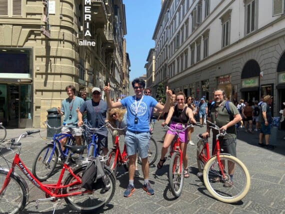 A guide in a blue t-shirt stands in front of his group of cyclists with two thumbs in the air.