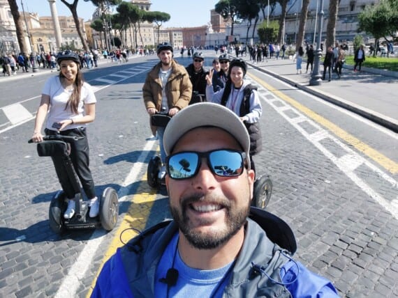 A guide wearing sunglasses takes a selfie with his group on Segways behind him.