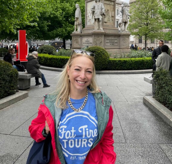 A woman in a blue t-shirt and red jacket smiling in front of a statue in Italy.