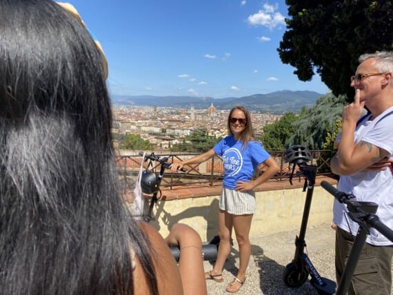 A women in a blue t-shirt and shorts stands next to an e-scooter at a lookout point over Florence, Italy.