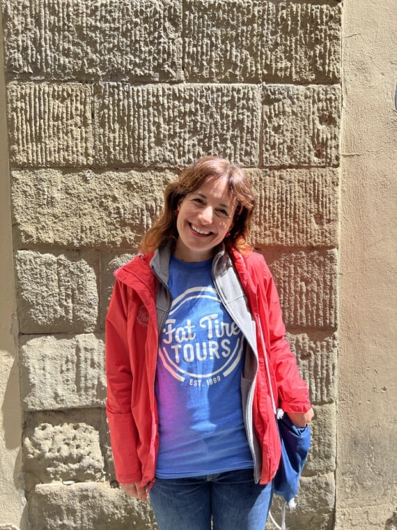 A smiling woman in a blue t-shirt and red jacket standing in front of a brick wall.