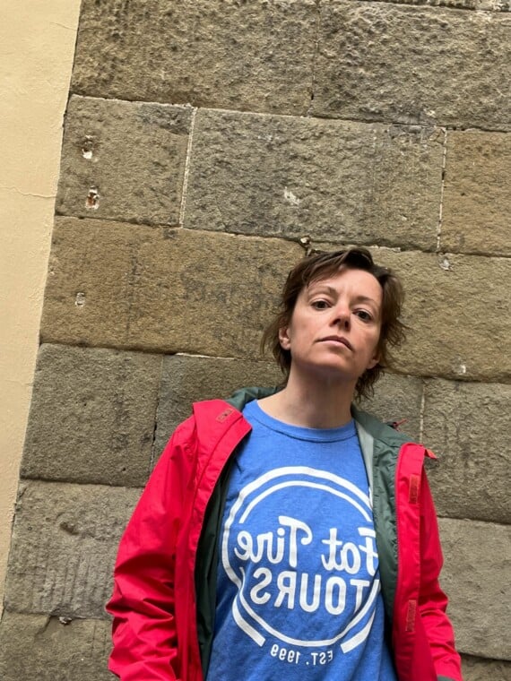 A woman in a blue t-shirt and red jacket leaning against a brick wall.