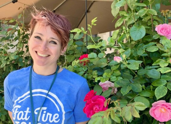 A smiling woman stands near a flowering bush in a blue t-shirt