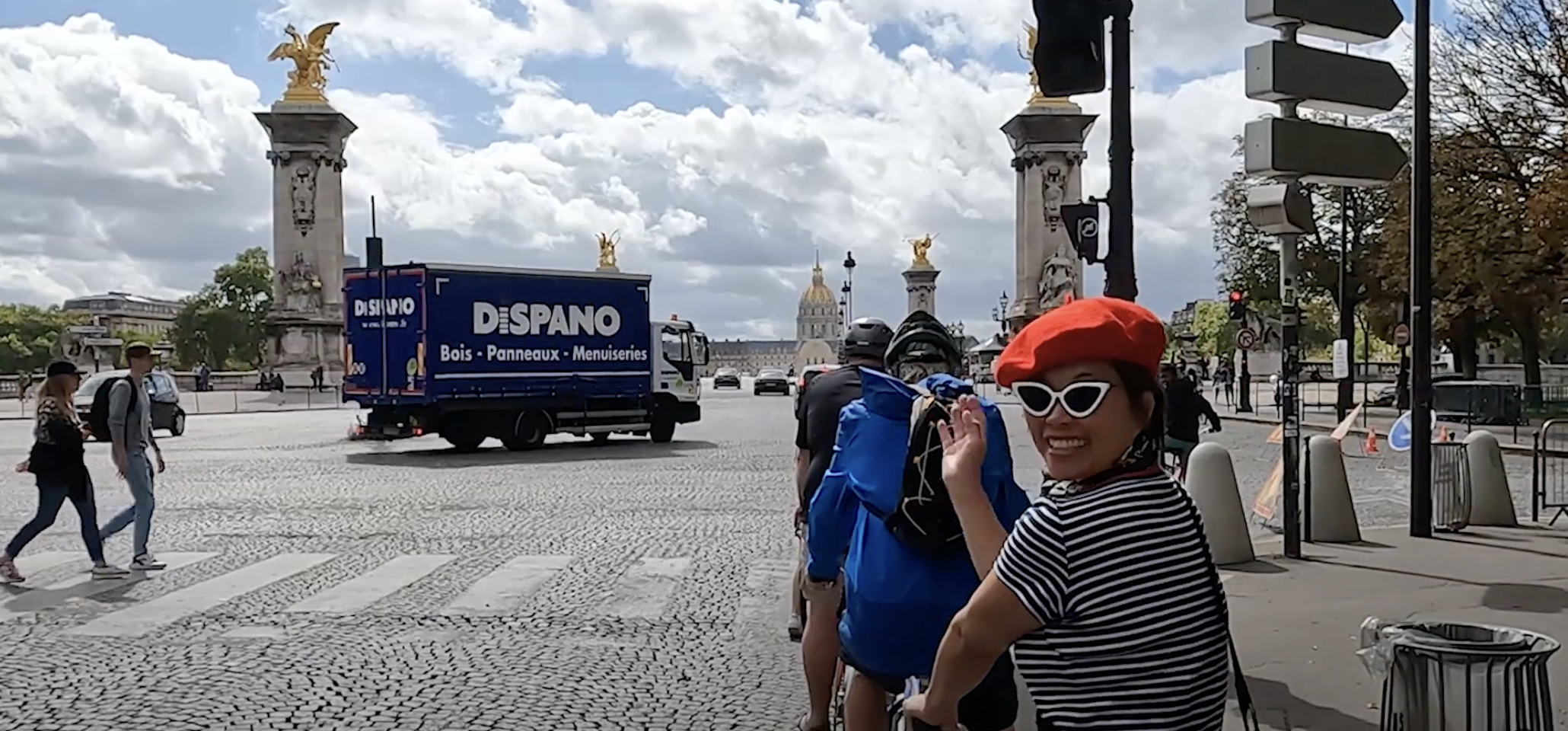 A woman in a striped shirt and red beret waves with the Alexandre III Bridge in the background