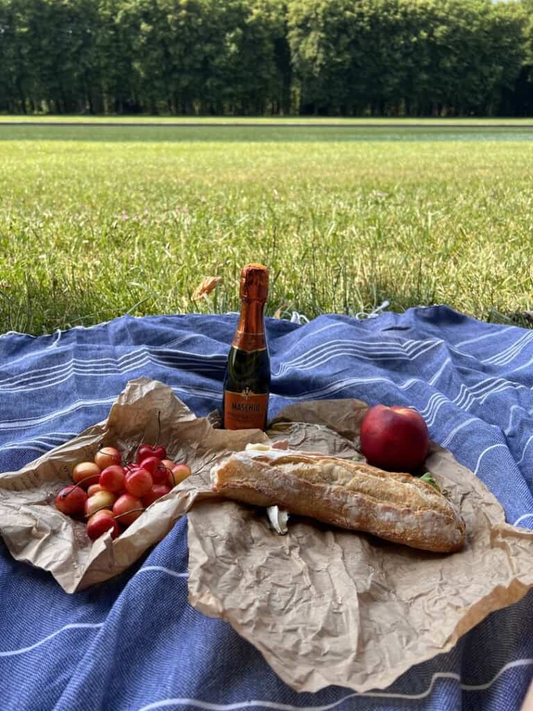 a sandwich, cherries, an apple, and a small bottle of champagne displayed on a picnic blanket