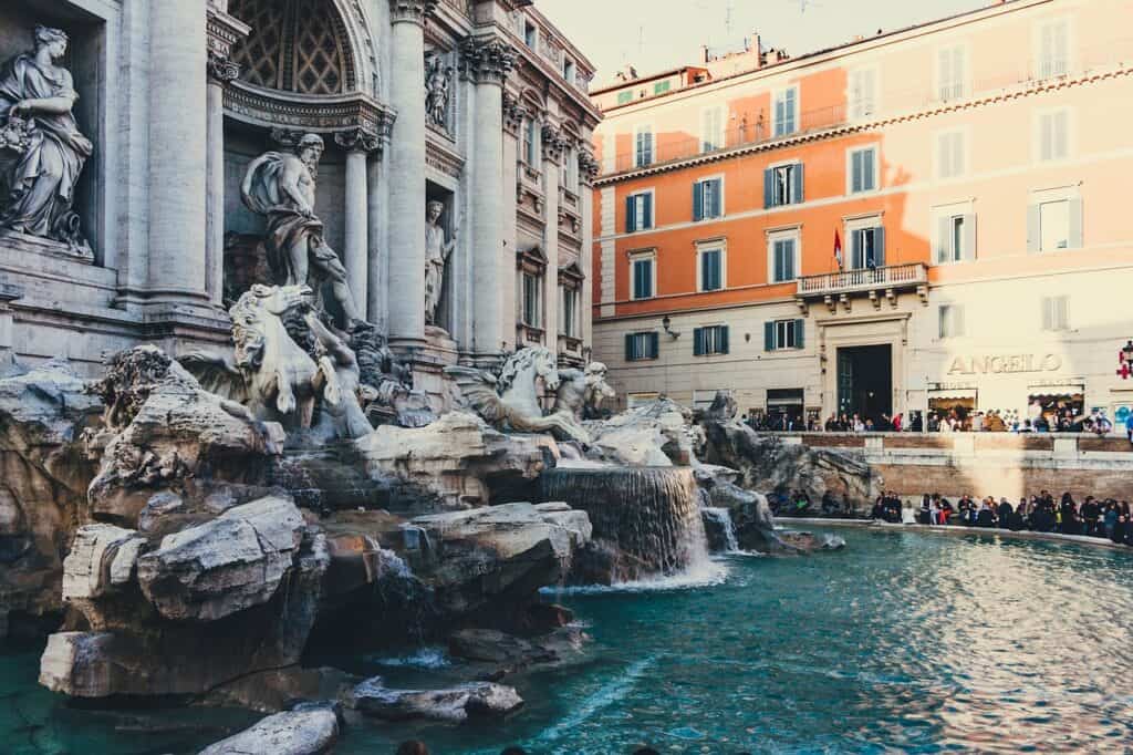 The Trevi Fountain is pictured with beautiful white statues and water pouring out of the fountain as visitors look on