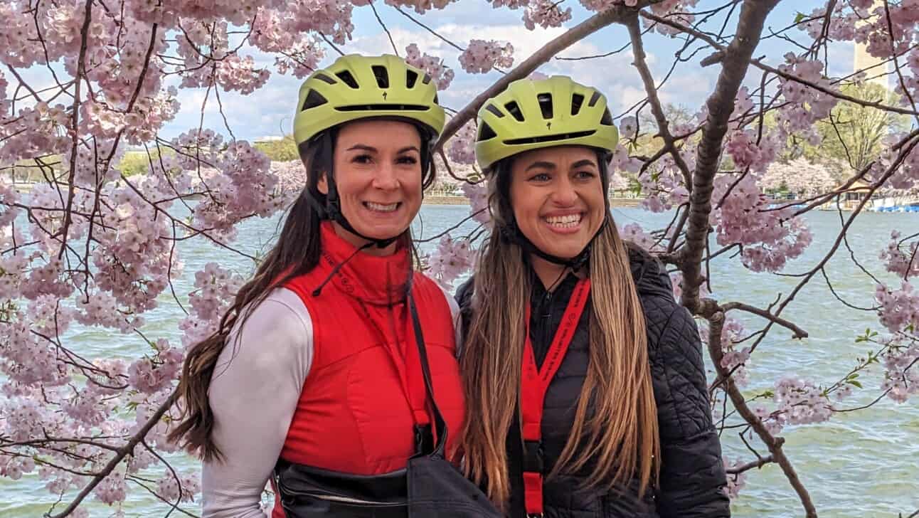 Two women with helmets on in front of the Cherry Blossoms in DC