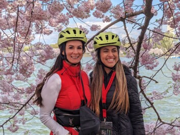 Two women with helmets on in front of the Cherry Blossoms in DC