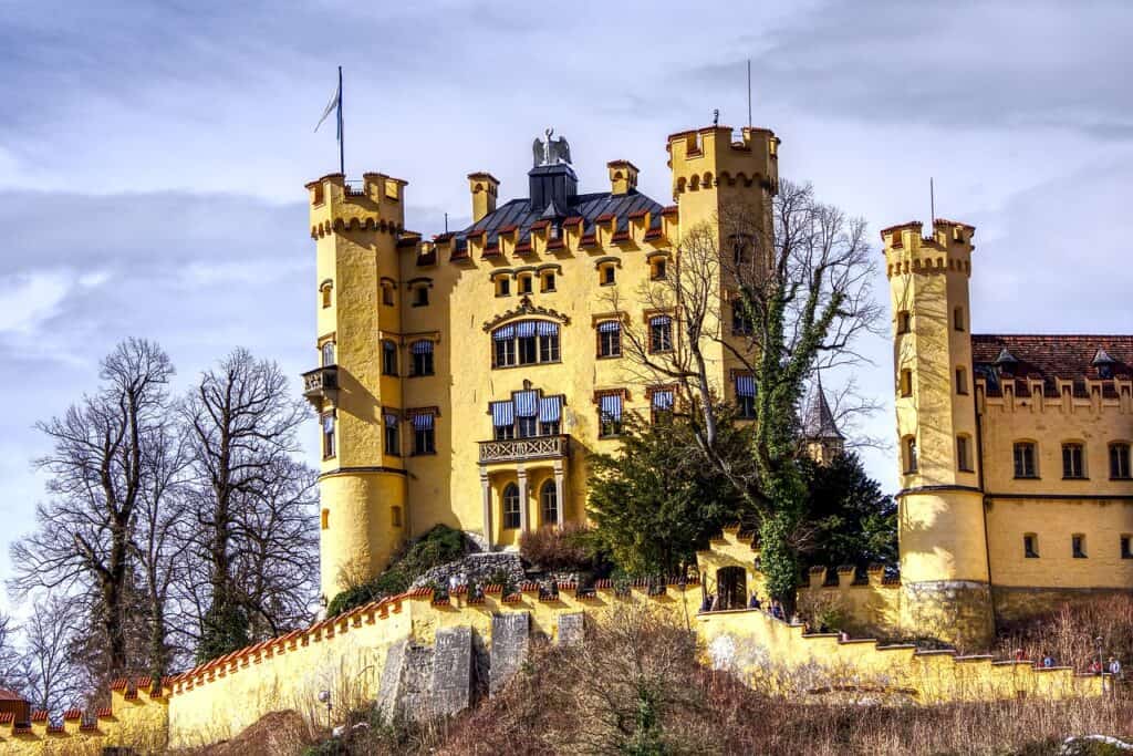 Hohenschwangau Castle is pictured sitting atop a hill. It has yellow-colored paint and many windows. 