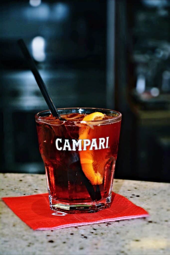 A Negroni cocktail in a Campari-branded glass with white lettering and a black straw