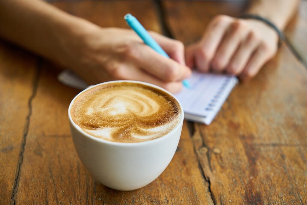 Latte pictured on a table as someone writes in a notebook in the background