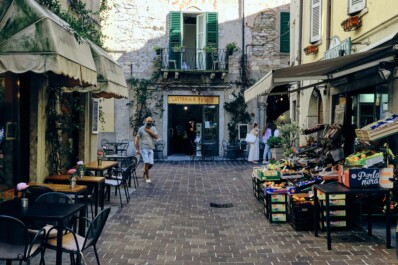 A man walking through Bellagio along Lake Como as a café spills out into the street and a fruit and vegetable stand is on display