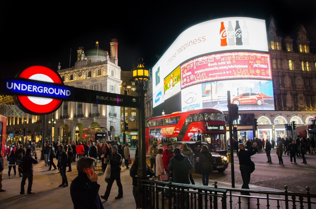 Piccadilly Circus at night in London with TV screens lit up and an Underground station sign in the foreground