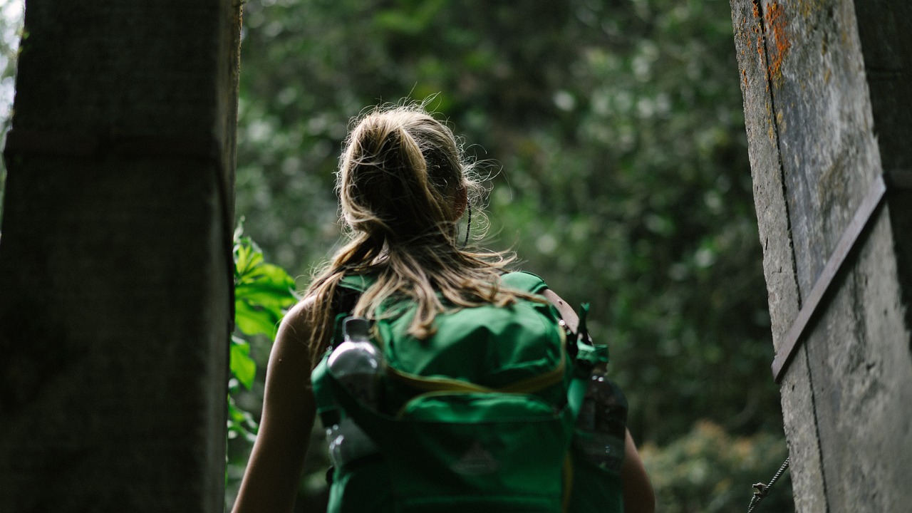 A blonde woman with a ponytail wears a green backpack and walks through the woods