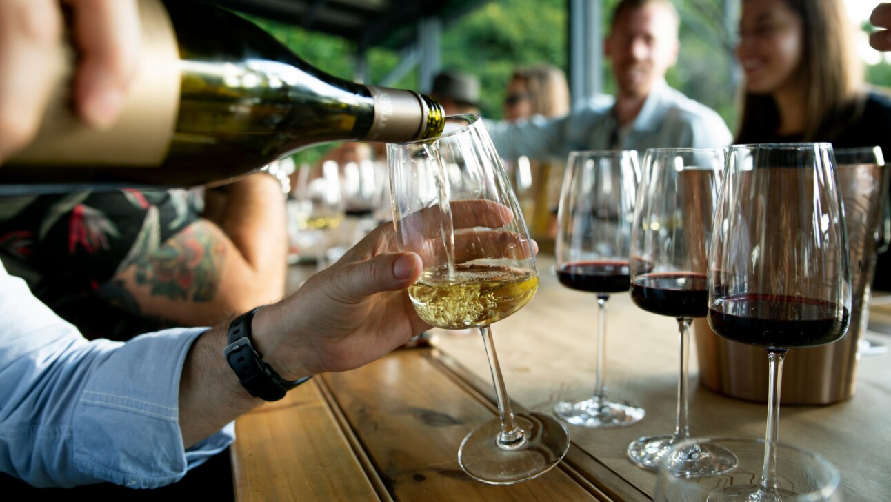 A man pours a glass of white white in front of three red wine glasses.